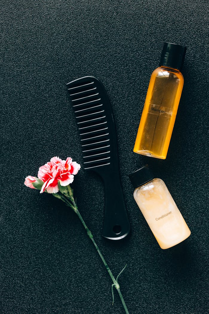 services-01 Flat lay of hair care essentials with a comb, bottles, and pink flower on dark background.